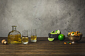 Tequila bottle and shot glasses with limes and small bowl of spicy rice crackers