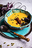 High angle of ceramic bowl with pumpkin cream soup with basil herb, rye bread and seeds served on blue plate with wooden spoon