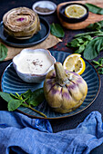 A steamed artichoke served with a bowl of yoghurt-mayonnaise dip on a dark blue plate