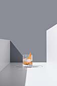 Crystal clear glass filled with fresh old fashioned cocktail garnished with orange and ice cubes placed on white surface between white walls against gray background