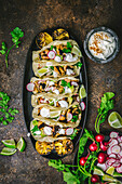 Arrangement of grilled chicken tacos on a dark platter with sliced radishes, limes, coriander, charred lemons and sour cream