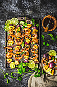 Skewers of grilled shrimp, pineapple and red onion on a baking tray and plate with glaze in bowl