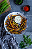 Fried okra fries in a white bowl, served with a white dipping sauce, whole okra pods, paprika and parsley leaves on the side.