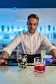 Male bartender serves refreshing whisky and Negroni cocktails in glasses while standing behind the counter and looking at blurred shelves of bottles