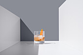 Crystal-clear glass filled with fresh old-fashioned cocktail, garnished with oranges and ice cubes on a white surface between white walls against a grey background