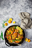 Chicken thighs in a cast-iron pan with fresh and roasted lemons, fennel and shallots on a grey background with lemon slices as garnish