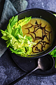 Healthy cream of celery soup in bowl with celery sticks on black plate with black spoon and napkin in front of blurred dark background