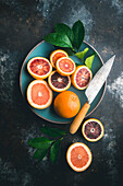 Blutorangen und rosa Grapefruit, halbiert und ganz, mit Zitrusblättern, in blauer Keramikschale und auf Tisch, mit Messer auf dunklem Hintergrund