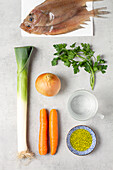 Top view of raw fish with carrot and onion with parsley herbs and spring onion stem with spices and cup as ingredients for Fumet placed on plain grey surface