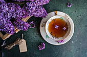 Tasty black tea in vintage white cup on mint green concrete table with aromatic lilac flowers