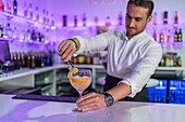 Focused male bartender garnishing a slice of orange on the rim of a cocktail glass with tongs while looking down and standing at the bar in front of blurry blue lights and bottles