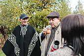 Happy mature men in traditional dress chat with an unrecognisable woman while drinking mate on a lawn in the park
