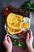 Hands holding a fork and a breakfast plate with French toast, fried egg and cherry tomatoes. Fresh mint leaves on the side