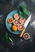 Blood oranges, halved and whole with citrus leaves, in blue ceramic bowl and on table, with knife on dark background