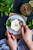 Woman dipping a leaf of a steamed artichoke into a yoghurt-mayonnaise sauce