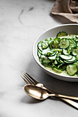 A bowl of German cucumber salad on a marble table with linen and gold cutlery