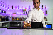 Male bartender standing behind the counter serving a clear glass of cold, refreshing whiskey with ice while looking down and working at the bar against a blurred purple background