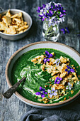 Green vegetable soup with croutons and edible violets in bowl with spoon and garnishes