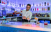 Focussed young male bartender looking down and pouring alcohol from bottle into jigger while preparing cocktail drinks and standing at counter
