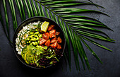 Lachs Poke Bowl Roher Fischsalat Asiatisches Trendgericht mit Sojabohnen, Edamame, Reis, Avocado und Salat in Schale auf tropischem Blatt und dunklem Hintergrund
