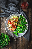 A plate of meatloaf, mashed potatoes and snap peas on a metal plate with peas and sauce on wood table