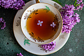 Top view of tasty black tea in vintage white cup on mint green concrete table with aromatic lilac flowers