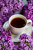 From above white cup of tea and beautiful lilac flowers around on grey concrete background