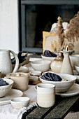 Rustikaler Tisch mit leerem, handgefertigtem Keramikgeschirr, weißen, rauen Schüsseln, Tellern, Tassen, Krug und Vase auf einem Leinentischtuch. Getrocknete Schilfblüten. Tageslicht Esszimmer