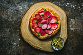 Round pastry tart with sliced beets, feta and mint leaves on wooden board with pistachios in bowl