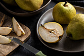 A halved pear, slices and a knife are placed on a chopping board. Fruit on a plate