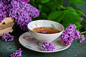 Vorderansicht eines leckeren schwarzen Tees in einer weißen Vintage-Tasse auf einem mintgrünen Betontisch mit aromatischen Fliederblüten