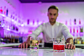 Male bartender serves refreshing whisky and Negroni cocktails in glasses while standing behind the counter and looking at blurred shelves of bottles