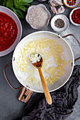 Diced onions being cooked in a white pot, chopped tomatoes, rice, spices, garlic cloves and basil leaves on the side.