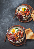Traditionelles komplettes englisches Frühstück mit Spiegeleiern, Würstchen, Bohnen, Pilzen, gegrillten Tomaten, Speck und Toasts auf grauen Tellern