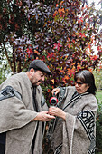 Serious mature couple in traditional Mapuche clothing with thermos and mug pouring hot yerba mate while standing in an autumn garden in Temuco
