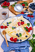 Crepes garnished with blueberries, raspberries, fresh mint leaves and powdered sugar served on a round plate.