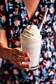 Hands holding a taro milkshake with whipped cream and straw against a floral dress background