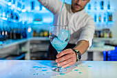 Male bartender holding a glass and looking down while pouring a blue coloured cocktail on ice into a transparent glass and spraying it on the bar counter in front of a blurred shelf of bottles
