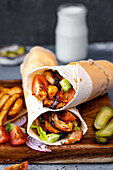Chicken doner kebab wraps on a wooden board accompanied by chips, pickles, tomatoes on the side and a glass of ayran yogurt drink behind them.