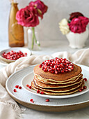 Stack of pancakes decorated with pomegranate and red flowers on a light background