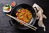 Udon noodles with prawns and vegetables in a wok pan on a black, burnt wood background