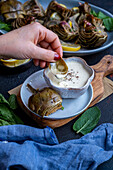 Woman hand holding a leaf of a steamed artichoke dipped into a yogurt and mayonnaise sauce.