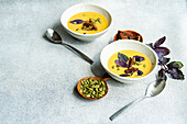 Schalen mit Kürbiscremesuppe mit Basilikumkraut, Roggenbrot und Kernen auf unscharfem grauem Hintergrund mit Blättern von oben