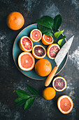 Blood oranges and pink grapefruit, halved and whole, with citrus leaves, in blue ceramic bowl and on table, with knife on dark background