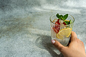 Front view of a cropped hand holding a summer cocktail glass with ice, mint, lemon and strawberry against a grey and white background