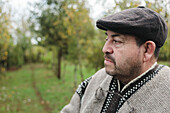 Nachdenklicher älterer Mann in traditioneller Mapuche-Kleidung und Baskenmütze schaut weg, während er vor einem verschwommenen Wald in Temuco steht