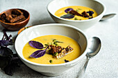 Bowls of pumpkin cream soup with basil herb, rye bread and seeds on a blurred grey background with leaves from above