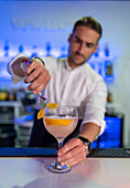 Focused male bartender using tongs to garnish a slice of orange on the rim of a cocktail glass while looking down and standing at the bar in front of blurred blue lights and bottles