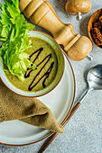 Top view of healthy celery cream soup in bowl with celery sticks on plate with napkin, spoon, jars and bowl with bread against grey background