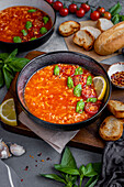 Tomato rice soup garnished with basil leaves, cherry tomatoes and a slice of lemon in a black bowl, bread slices, basil leaves and another bowl of soup behind it.
